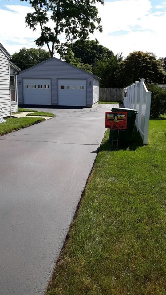 A paved driveway leads to a light grey two-car garage, with a white vinyl fence and a trash can on the right side.