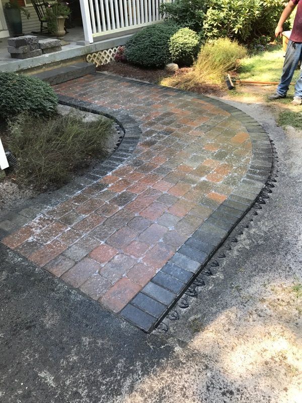 A curved brick paver walkway with a dark grey border leads to a porch with white railings, set against a yard with greenery.
