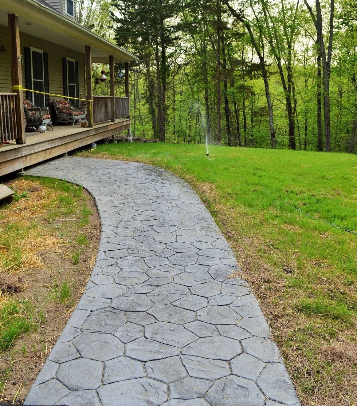 A gray, flagstone-patterned concrete walkway leads to the covered porch of a house near a wooded area.