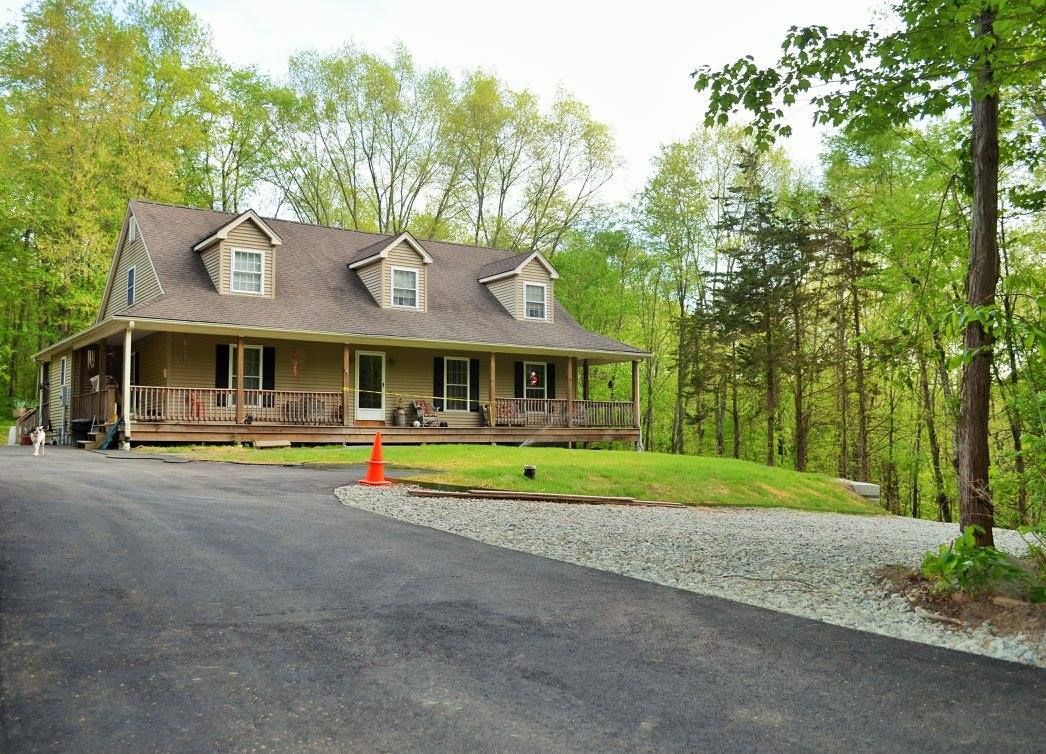 A tan house with dormer windows and a covered front porch, surrounded by lush trees, viewed from a gravel driveway.