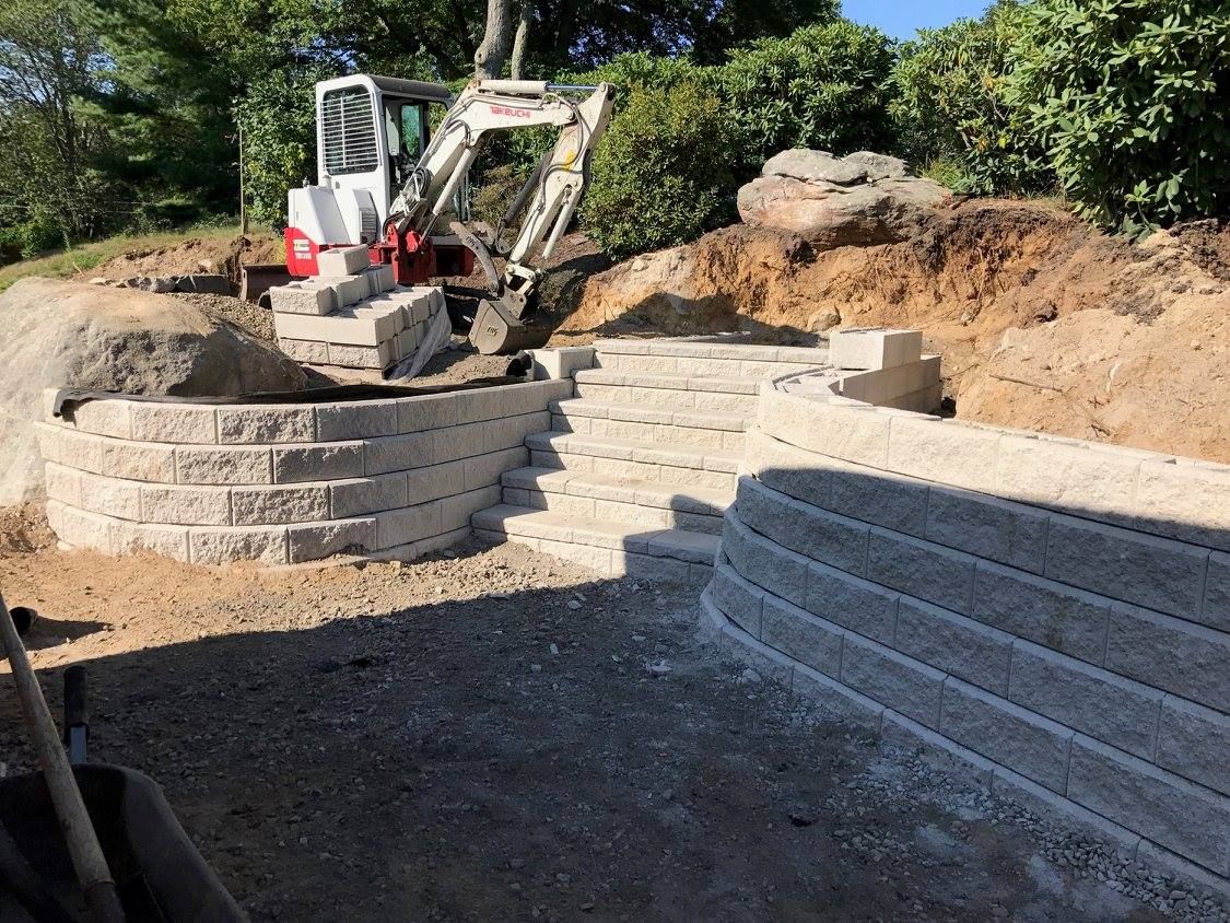 A small white excavator parked on a dirt slope above a newly constructed set of light gray concrete retaining walls.