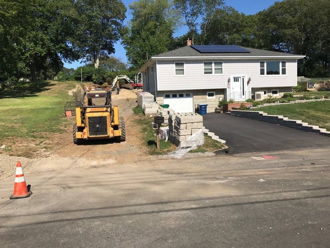 A construction vehicle sits on a dirt lot next to a light-colored house with a newly paved driveway and a traffic cone.