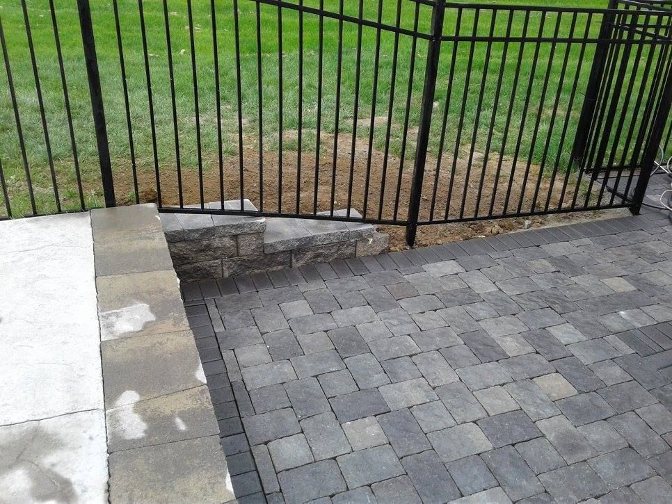 A gray paver patio meets a small stone retaining wall and a black metal fence bordering a patch of grass.