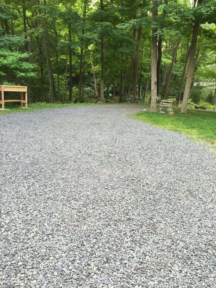 A gravel driveway curves through a wooded area with tall trees and green grass alongside it.