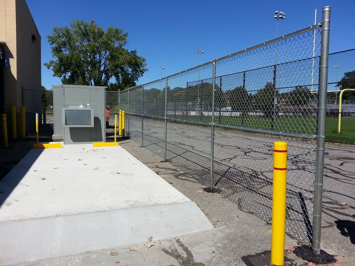 Concrete pad with a gray metal cabinet, enclosed by a chain-link fence, with yellow bollards in an outdoor parking lot.