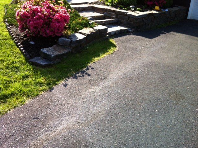 A vibrant pink azalea bush sits in a landscaped garden bed next to stone steps leading to a dark asphalt driveway.