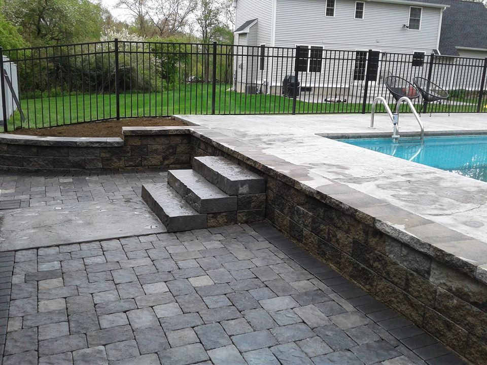 A stone patio with steps leading up to a swimming pool deck, enclosed by a black metal fence in a backyard.