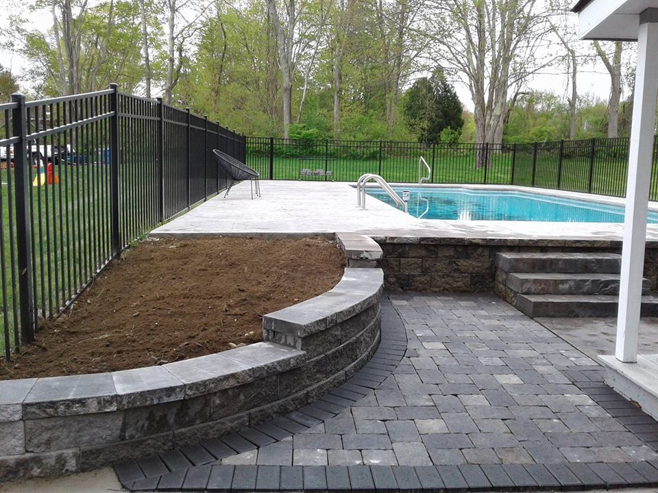 A landscaped pool area featuring a stone retaining wall, paved patio, steps leading to the pool, and a black metal fence.
