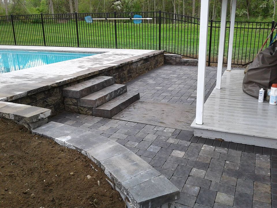 A swimming pool with stone steps leading down to a gray paver patio and an adjacent porch under a black metal fence.