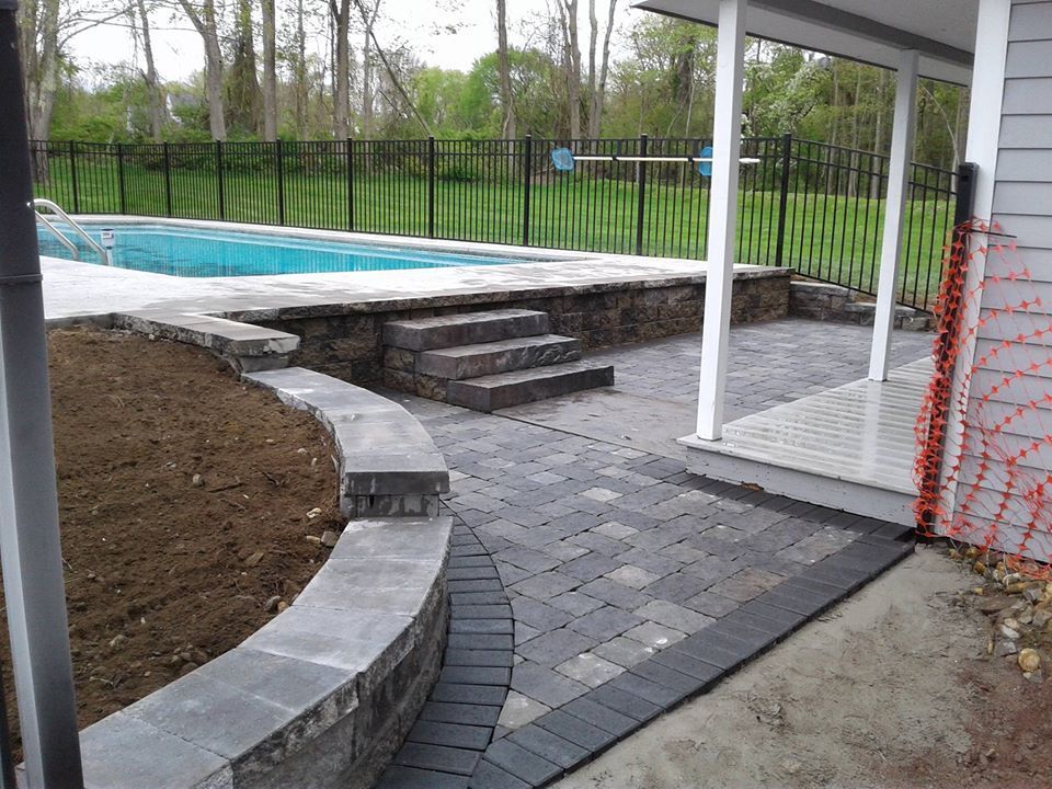 A stone patio with a curved retaining wall and stairs leading up to an in-ground pool area next to a house.