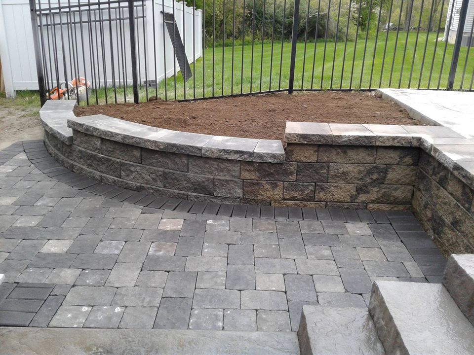 A raised garden bed with stone retaining walls built next to a gray paver patio and concrete steps in a fenced backyard.