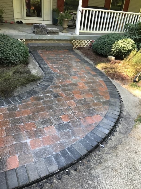 A paved walkway leading to a house porch, featuring multi-colored bricks bordered by dark gray stones and edge restraints.