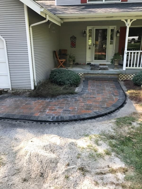 A curved brick walkway leads to a front porch and white front door of a house with light gray siding.