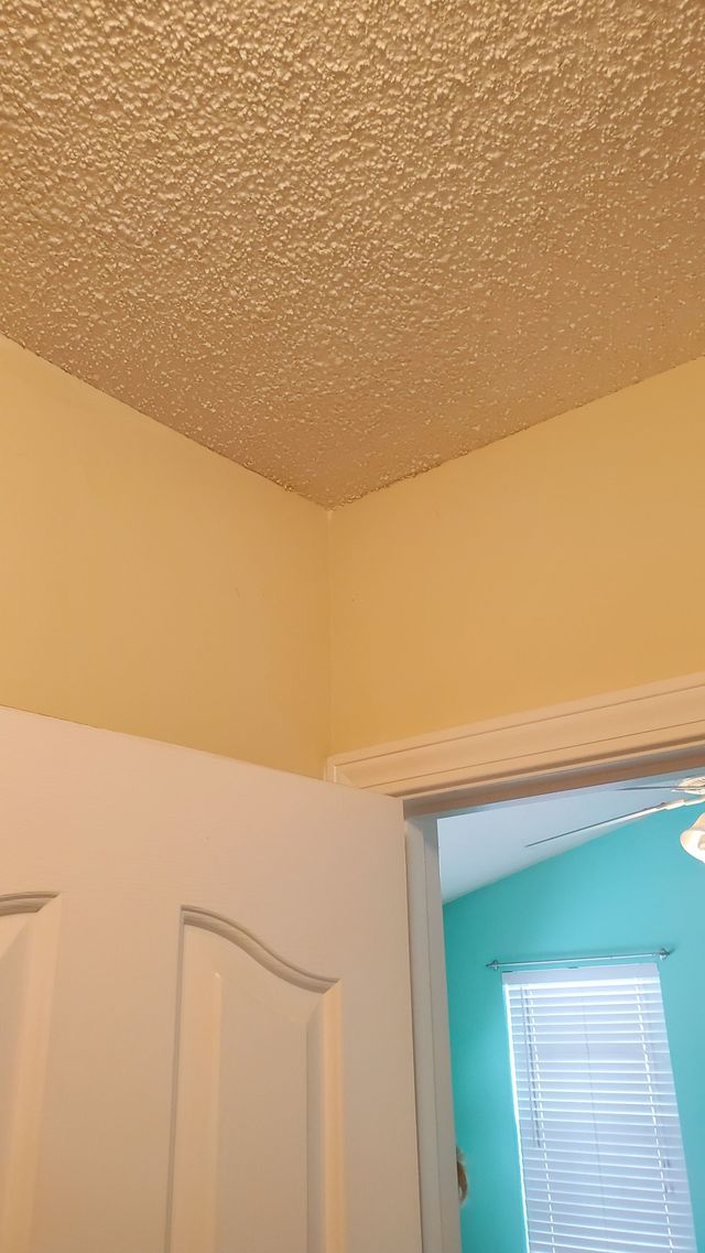 Corner of a beige room with a textured ceiling and open white door, showing a blue room beyond