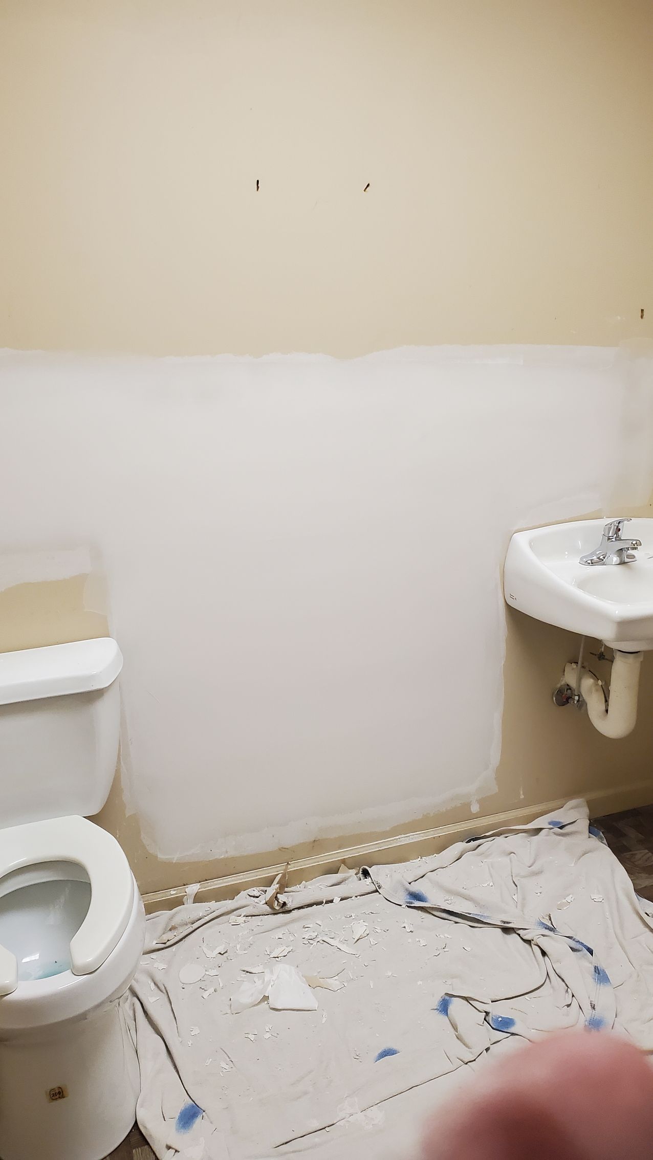 Small bathroom under renovation with toilet, sink, bare walls, and drop cloths on the floor.
