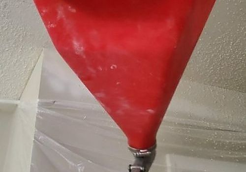 Red funnel-shaped bag with a metal valve against a light wall and translucent plastic sheeting.