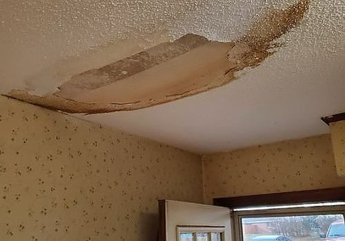 Water-damaged ceiling with peeling plaster above a window in a small room
