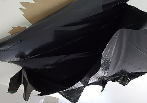 Black and gray torn plastic sheeting hanging against a white wall, with a strip of brown material behind it