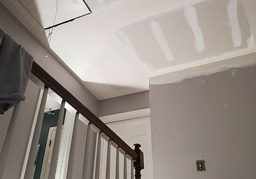 Stairwell with unfinished drywall ceiling and walls, showing taped patches and a wooden railing.