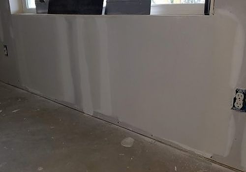 White interior wall with exposed electrical outlet and unfinished drywall over concrete floor.