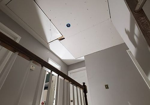 Stairwell ceiling with a large hole and dangling panel above a wooden railing