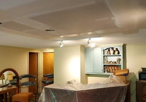 Living room with plastic-covered furniture, ceiling patchwork, and a built-in cabinet with dishes.