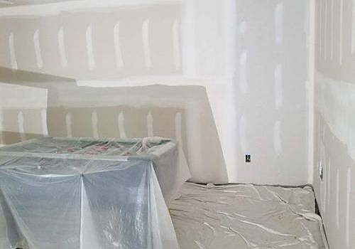 Unfinished room with taped drywall, covered furniture, and plastic sheeting on the floor