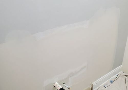 Partially painted white wall with patchy repair spots and a baseboard at the bottom