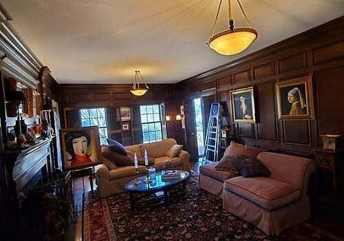 Cozy wood-paneled living room with sofas, chandelier, rugs, and framed portraits by the windows
