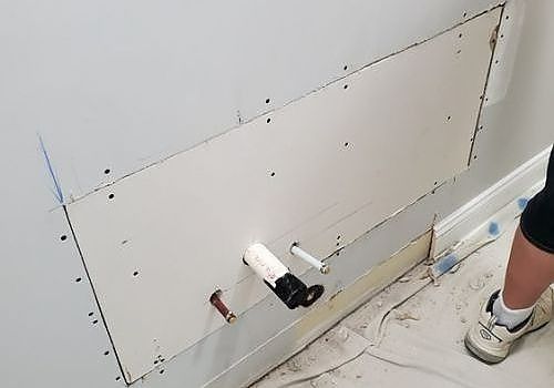 Bathroom wall under renovation with a drywall patch, exposed pipes, and a worker’s legs at right
