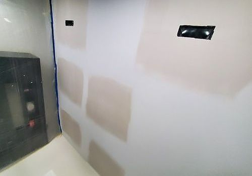 Unfinished white wall with patching, a small black window, and a dark cabinet in the corner.
