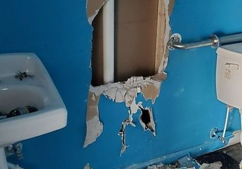 Damaged blue bathroom wall with a large hole exposing pipes and torn drywall near a sink and outlet.