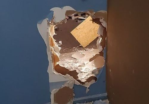 Damaged wall with a large hole exposing insulation and drywall, next to a blue wall and brown panel