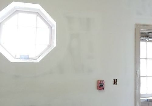 Empty white interior wall with an octagonal window, wall outlet, and a small red fire alarm box
