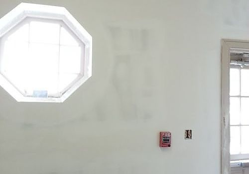 White wall with an octagonal window, small red alarm panel, and a doorway on the right.