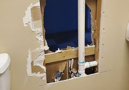 Damaged bathroom wall with exposed blue insulation and plumbing pipes behind torn drywall.