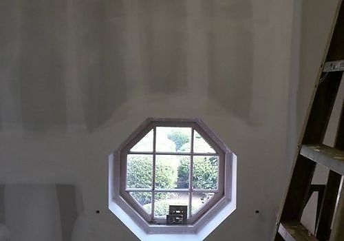 Octagonal window with white curtain and greenery outside, viewed from inside an unfinished room