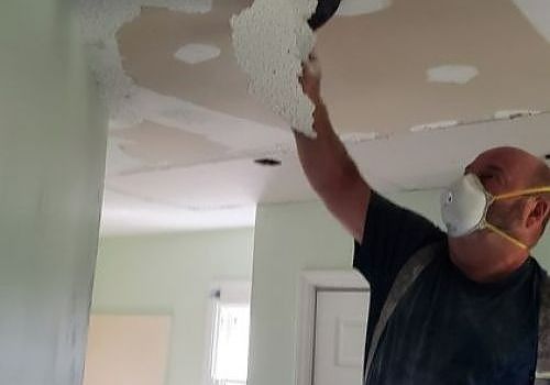 Man wearing a mask scraping peeling ceiling paint in a room under renovation