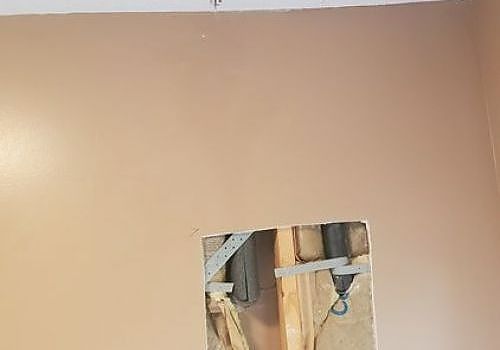 Beige wall with a rectangular hole exposing wooden framing and a hanging cable