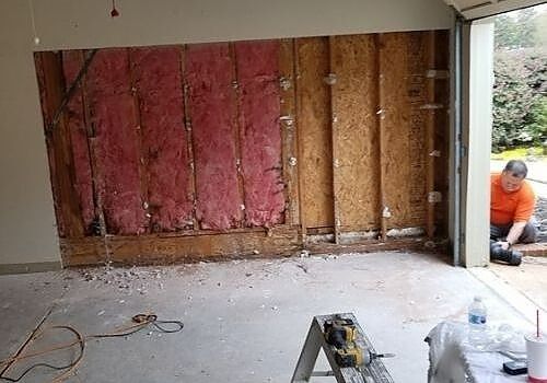 Partially gutted room with exposed wall insulation and a worker visible outside the doorway