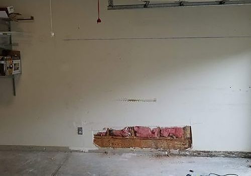 Empty room with white wall, exposed damage near the floor, and concrete floor with debris