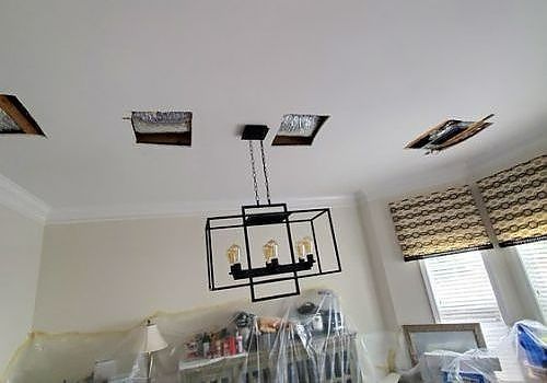 Living room with ceiling holes, a hanging black light fixture, and covered furniture during renovation