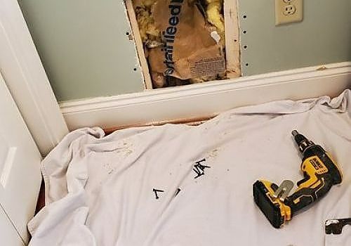 Bedroom wall with hole in drywall, white bedding, and a yellow cordless drill on the bed