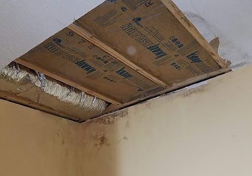 Damaged ceiling with a large hole exposing insulation and wooden beams above a stained wall.
