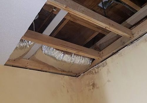 Ceiling opening with exposed wooden beams and silver ducting above a beige wall