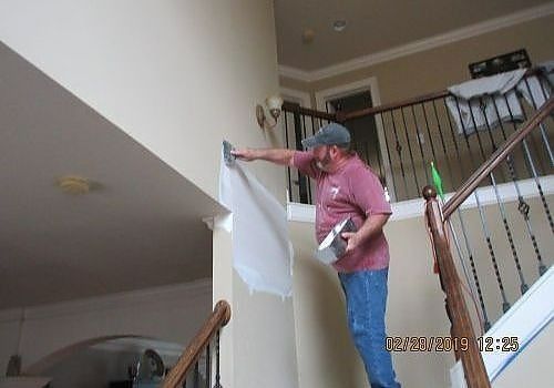 Person painting a stairwell wall with a roller, wearing a pink shirt and blue jeans.