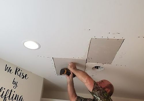 Person using a drill to install a ceiling vent panel in a room with a recessed light and wall decal.