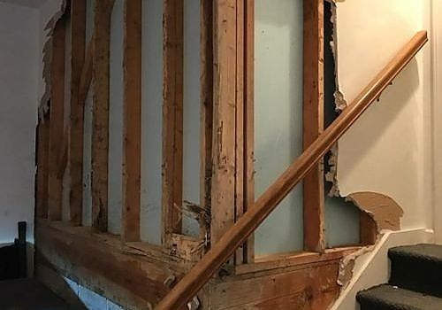 Damaged interior wall with exposed studs beside a wooden stair railing and carpeted steps