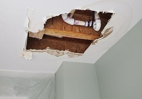 Damaged ceiling with a large hole exposing wooden beams and pipes in a room