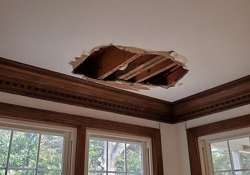 Ceiling hole exposing wooden beams above windows in a room with decorative trim.